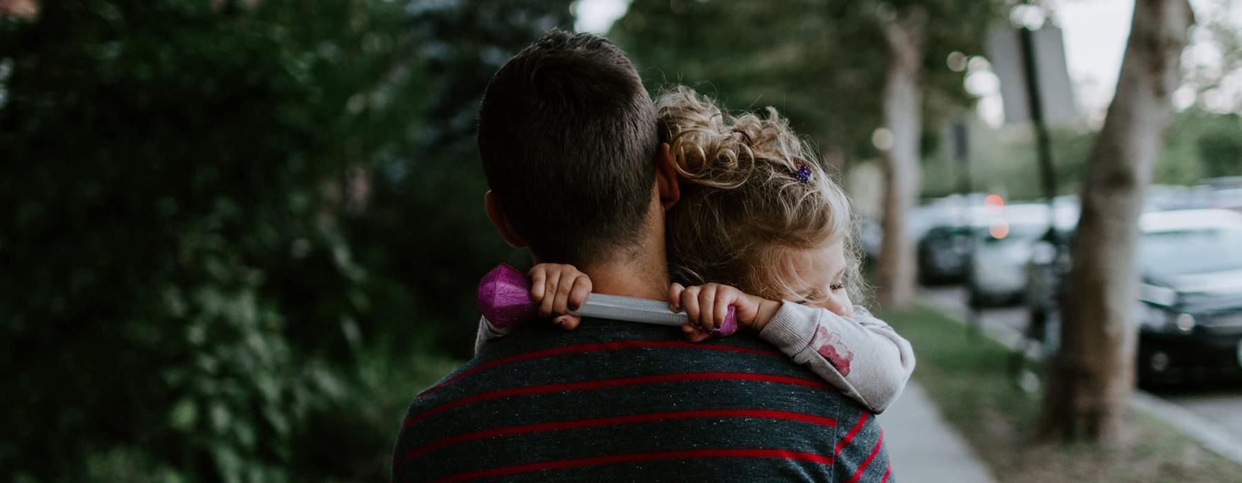 tired little girl rests her head on her father's shoulder as he carries her down the city sidewalk